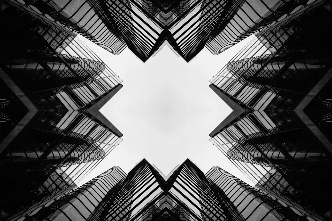 Symmetrical black and white cityscape looking up at mirrored glass office towers forming an X-shaped frame.