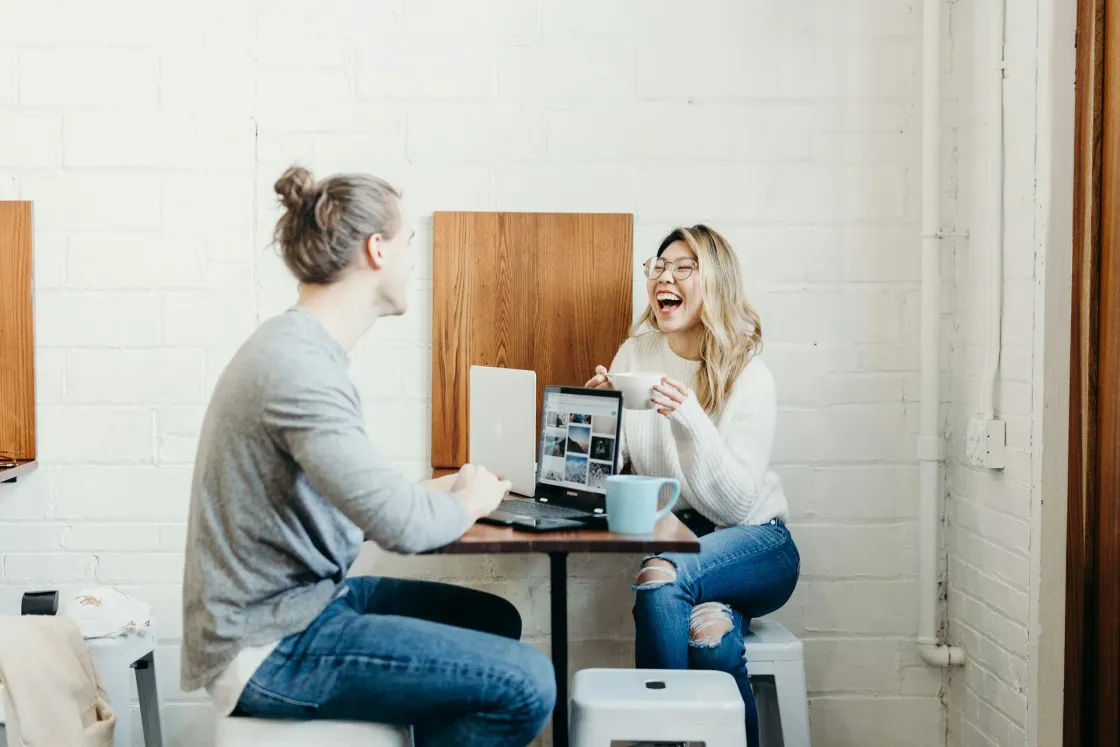 Two colleagues laughing over coffee and laptops in a bright workspace - capturing genuine friendship and connection at work.