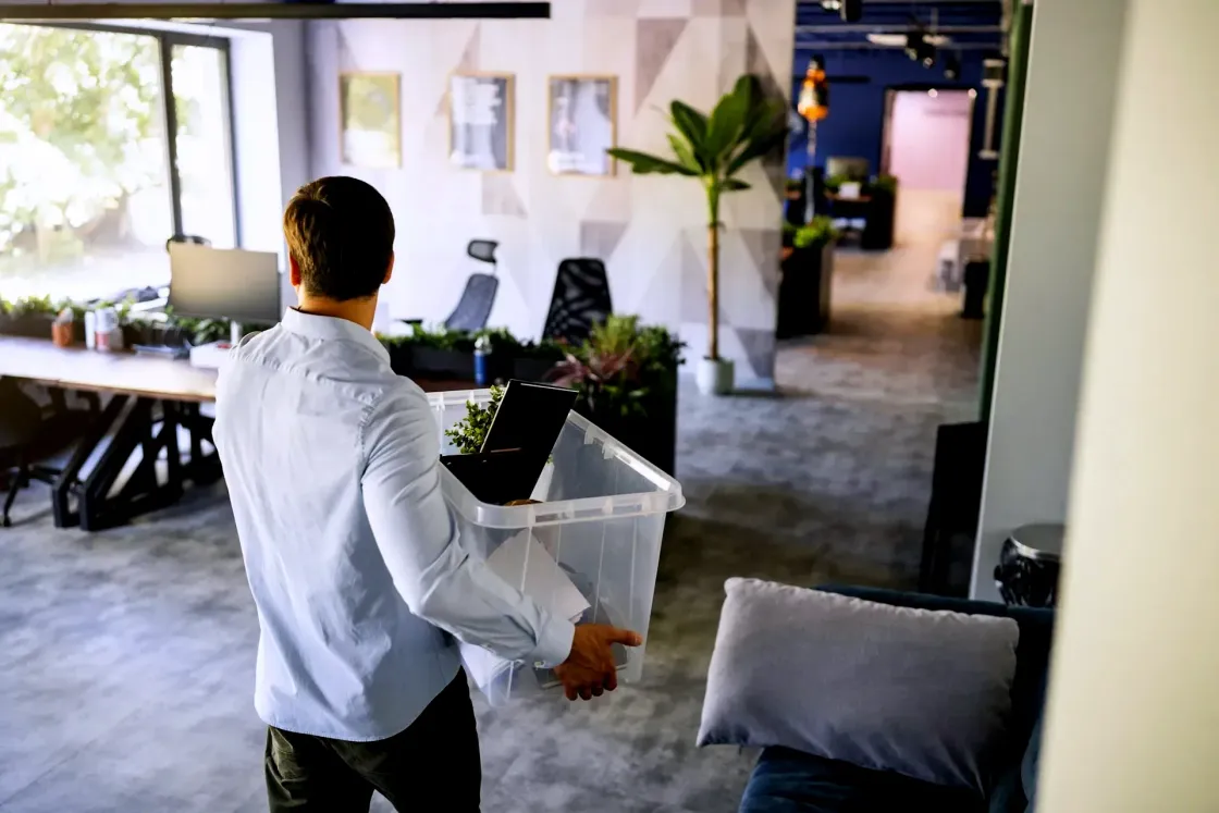 Employee carrying a clear plastic box of personal belongings through a modern open plan office after being laid off.