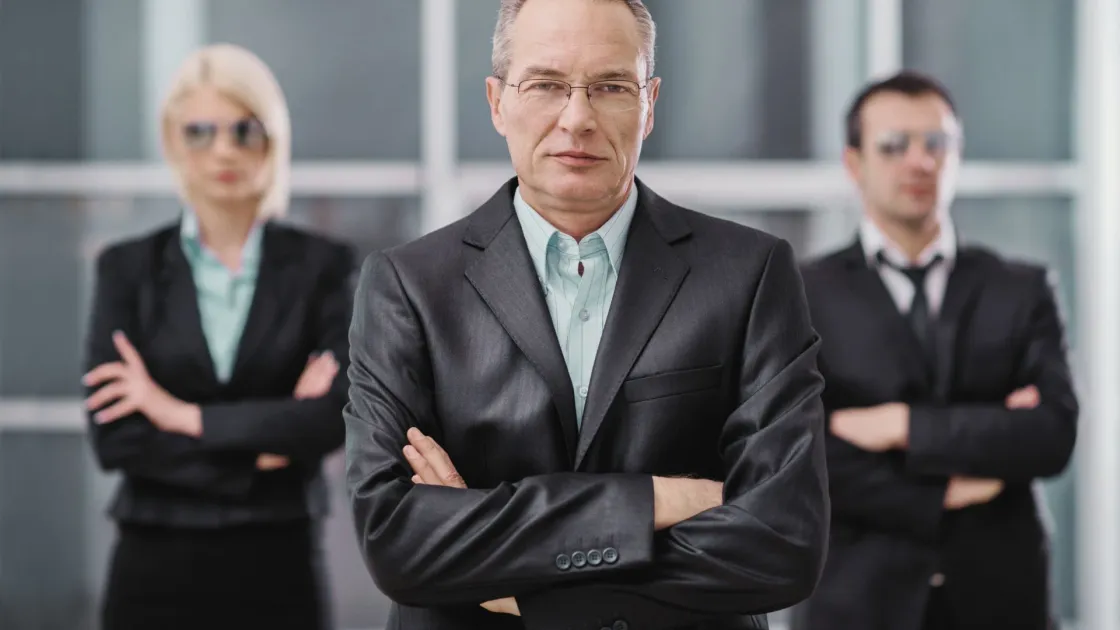 Stern executive in a dark suit with arms crossed, two colleagues blurred behind, symbolizing a top-down “agree or quit” workplace ultimatum.