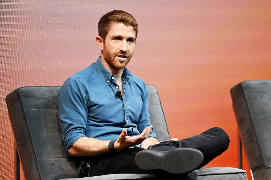 Bearded tech ethicist in a denim shirt speaking on stage, mid-sentence, during a discussion about artificial intelligence and the future of work.