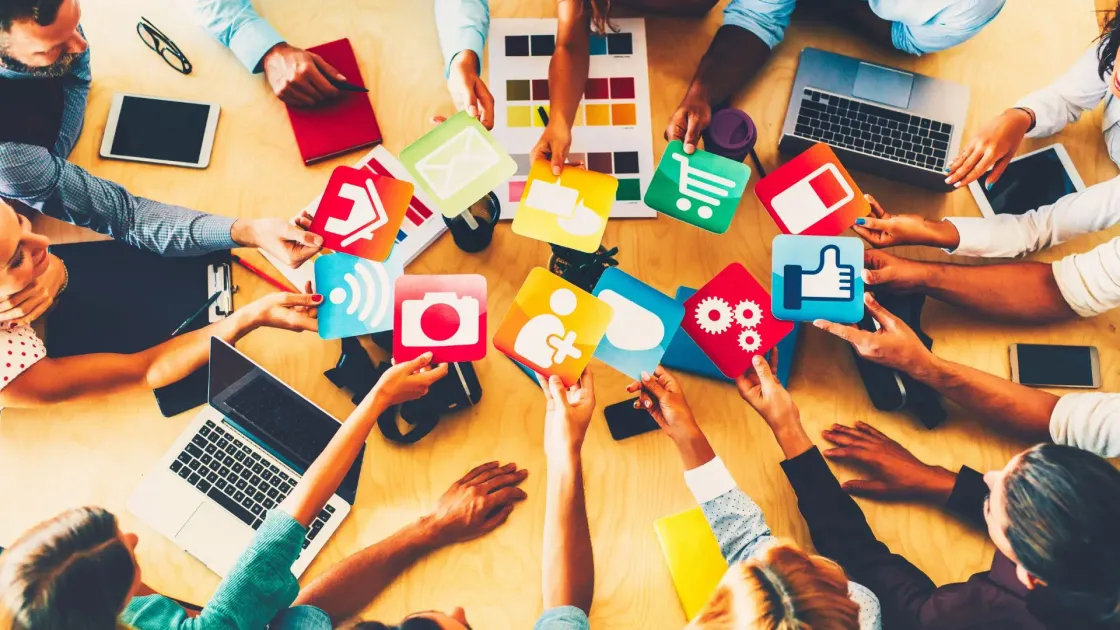 Overhead view of a team around a table with laptops and colourful social media icons, overlaid with bold white text reading “RECRUITING IS MARKETING”