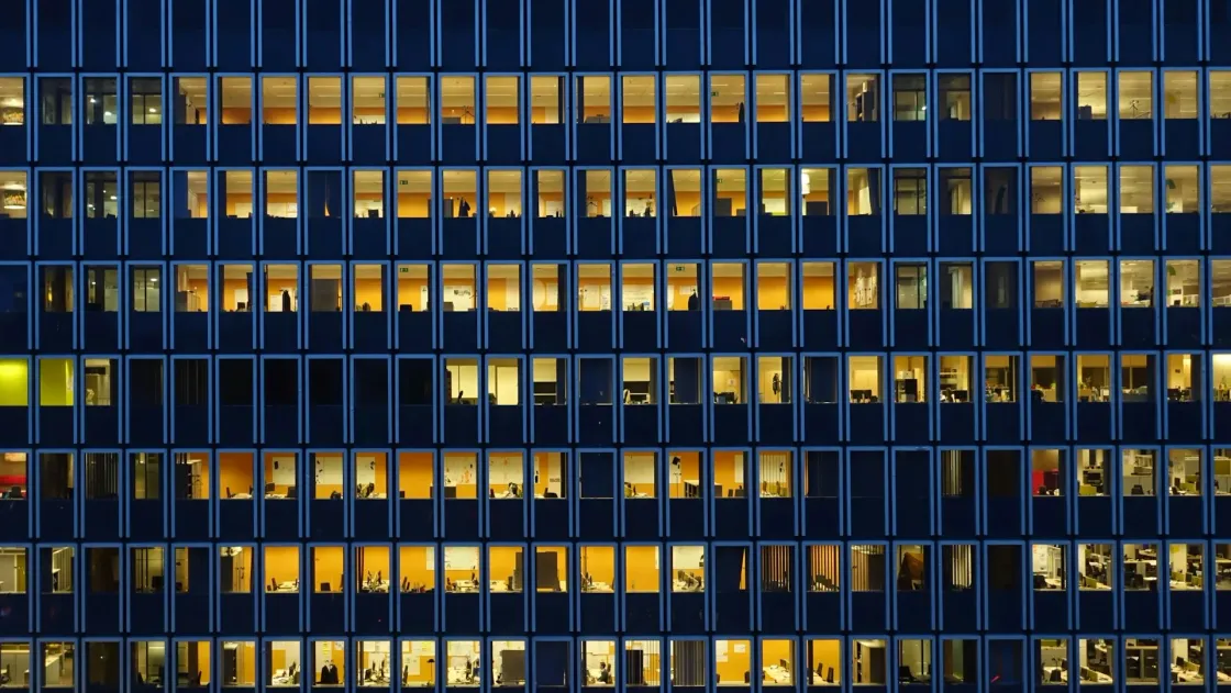Office building at night with rows of lit windows, symbolising corporate growth with lean headcount and fewer people inside.