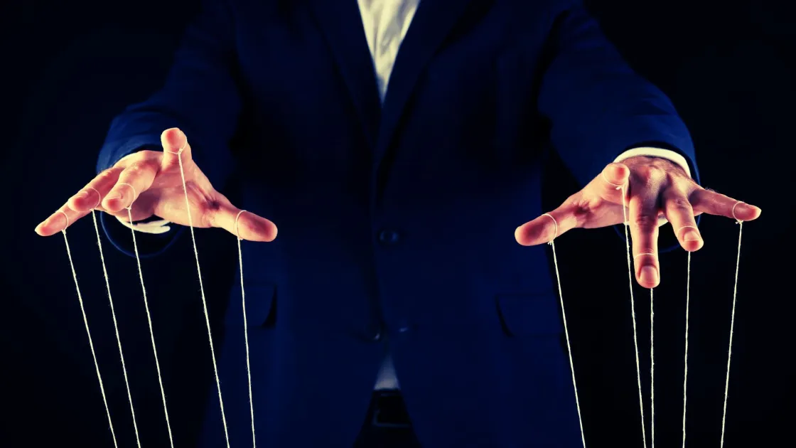 Person in a suit holding marionette strings, symbolising workplace control, monitoring, and management influence over employees.