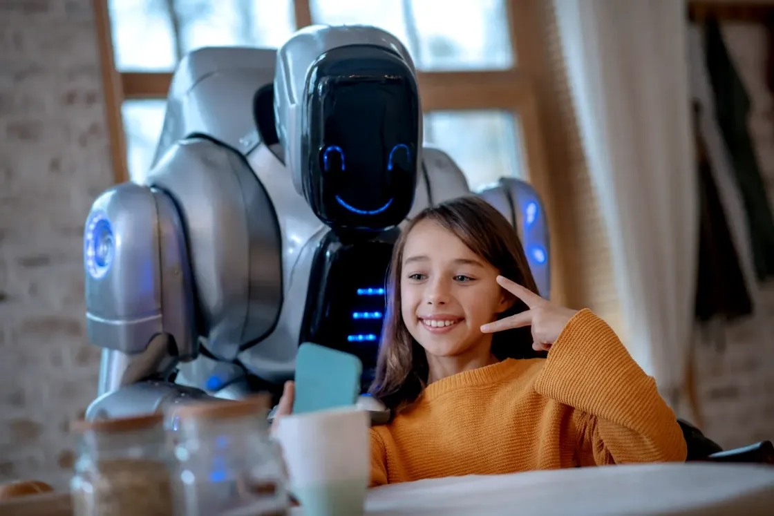 A smiling child in a mustard jumper takes a selfie in front of a humanoid robot at home.