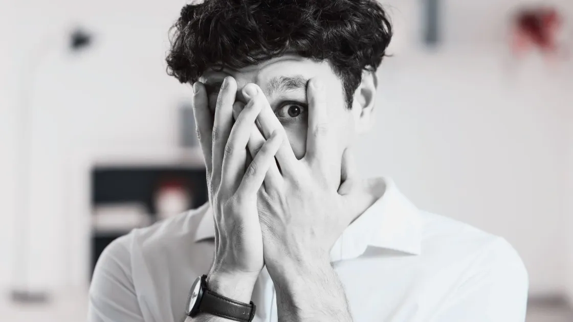 A young person covering most of their face with their hands, one eye visible, conveying anxiety, overwhelm, or apprehension in response to workplace change.
