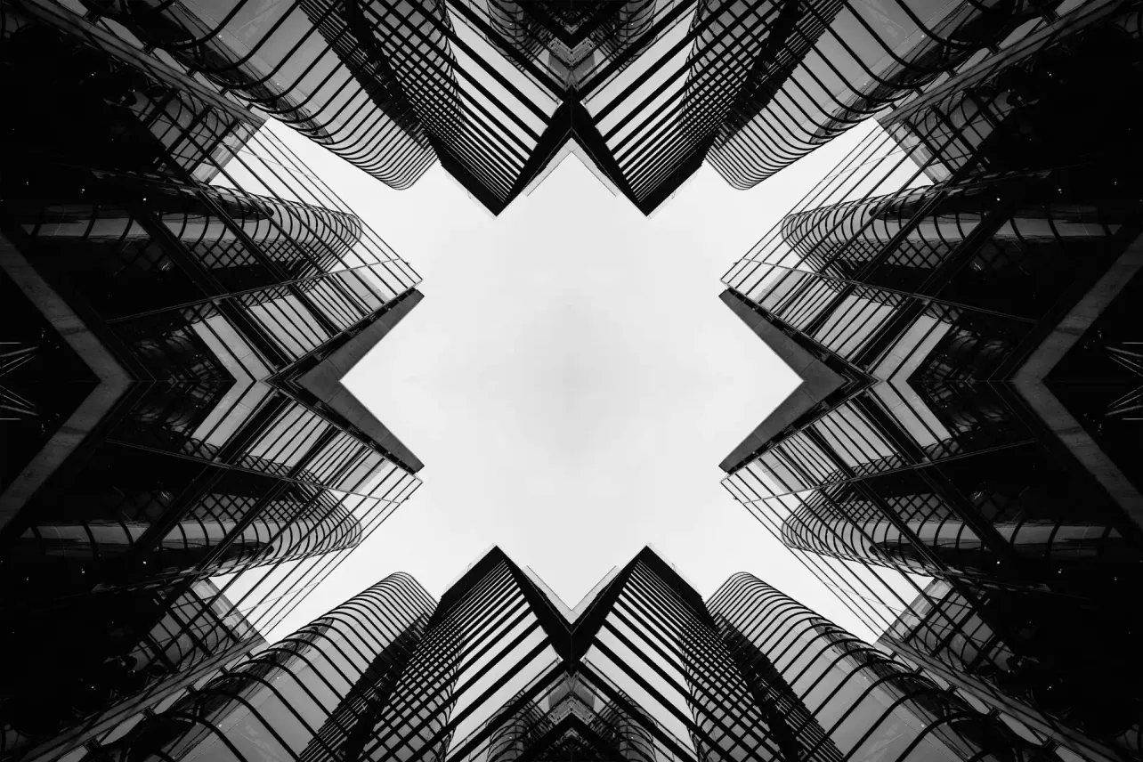 Symmetrical black and white cityscape looking up at mirrored glass office towers forming an X-shaped frame.