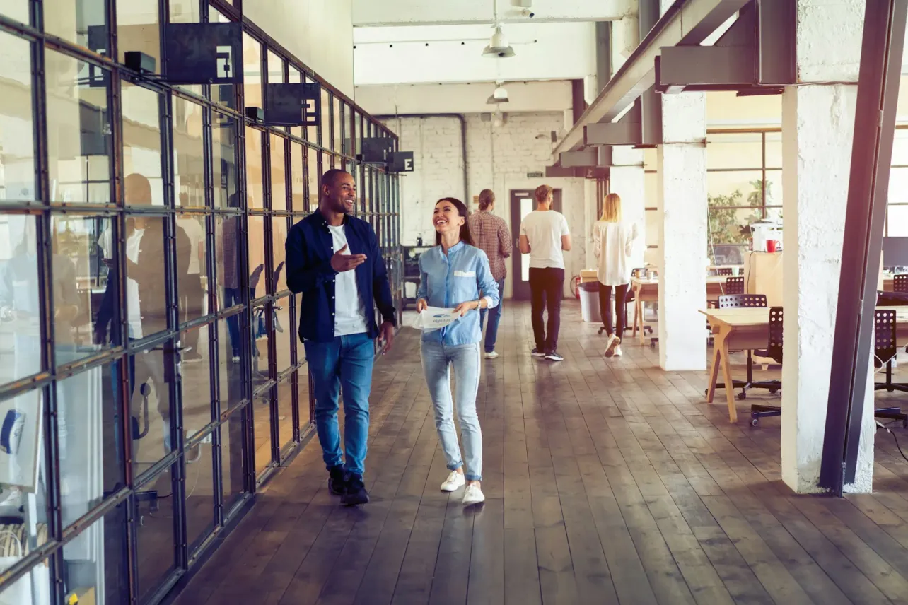 Two colleagues walk and talk down a bright open plan office corridor with glass meeting rooms on one side and desks on the other, while other coworkers move and work in the background.