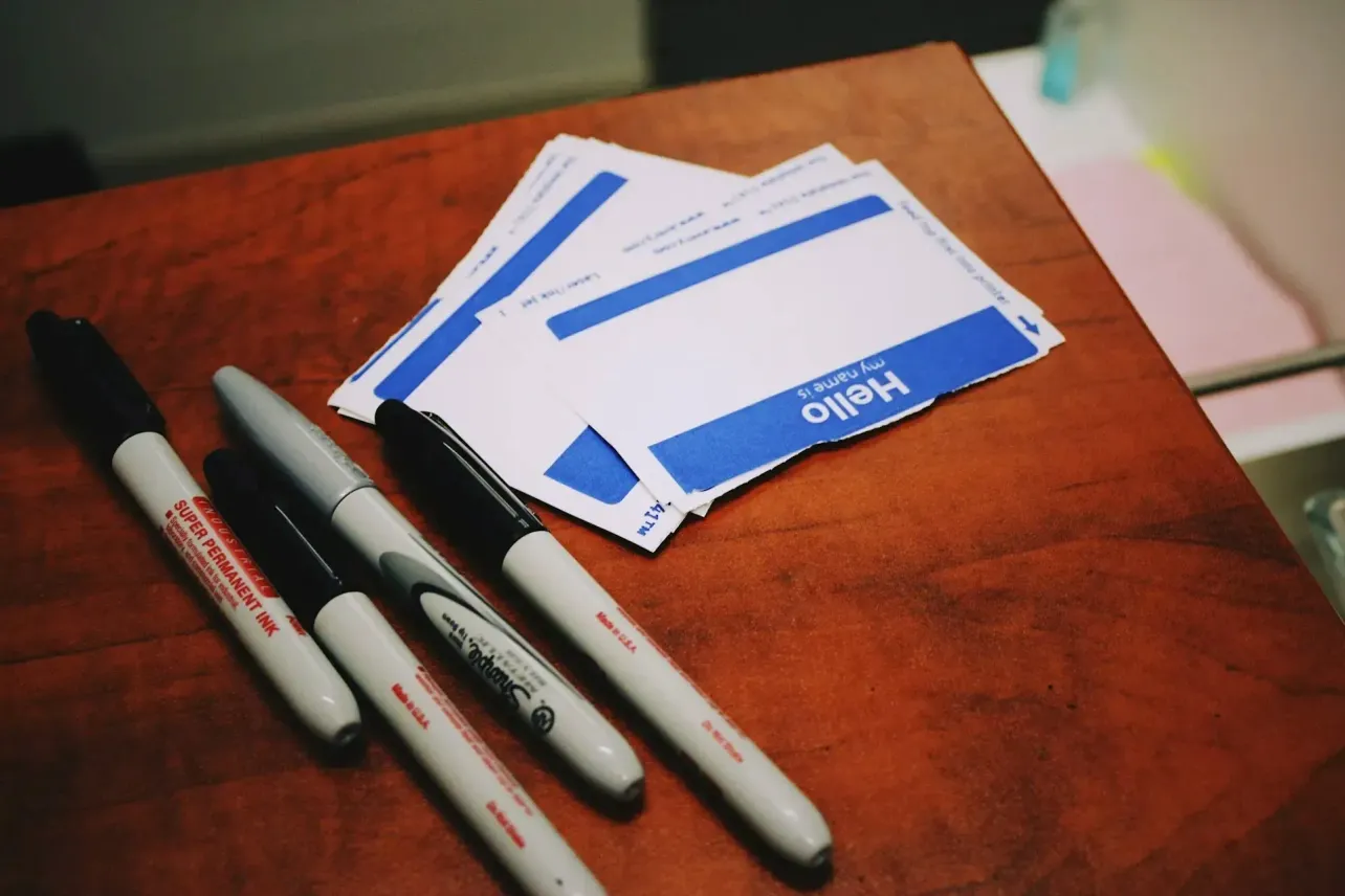 A stack of blank “Hello, my name is” stickers beside permanent markers on a wooden desk, symbolizing identity, self-definition, and new beginnings.