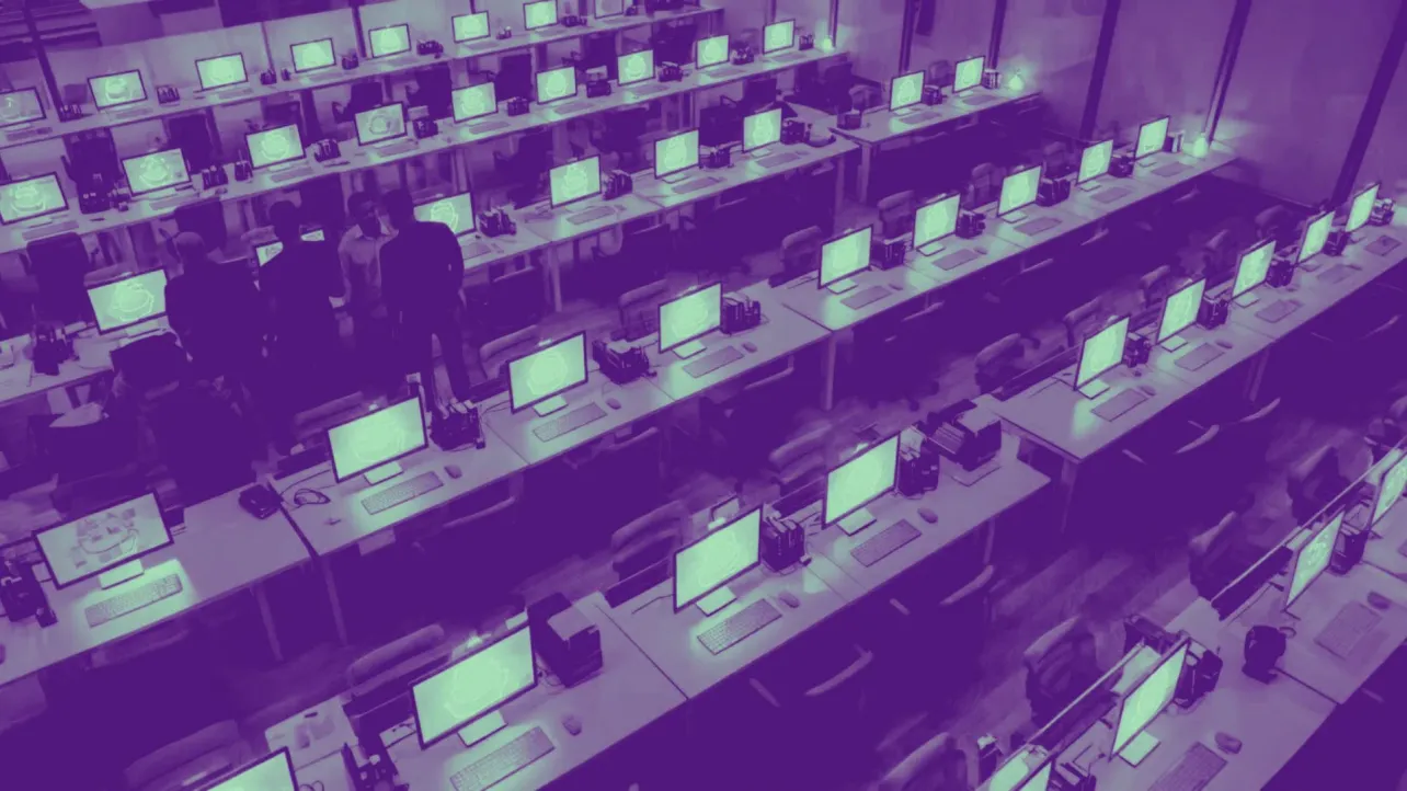 Rows of glowing computer screens in a purple tinted open plan office, with most chairs empty and a small group of workers standing together among the desks.