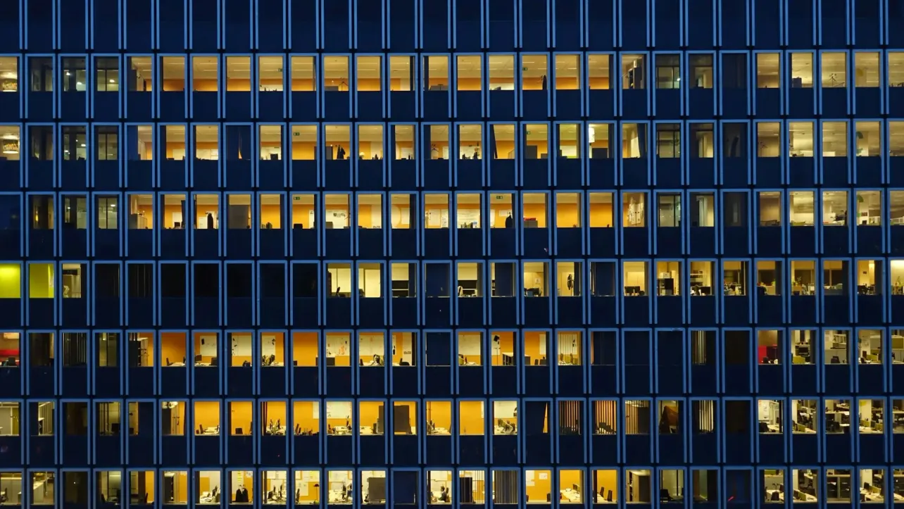 Office building at night with rows of lit windows, symbolising corporate growth with lean headcount and fewer people inside.