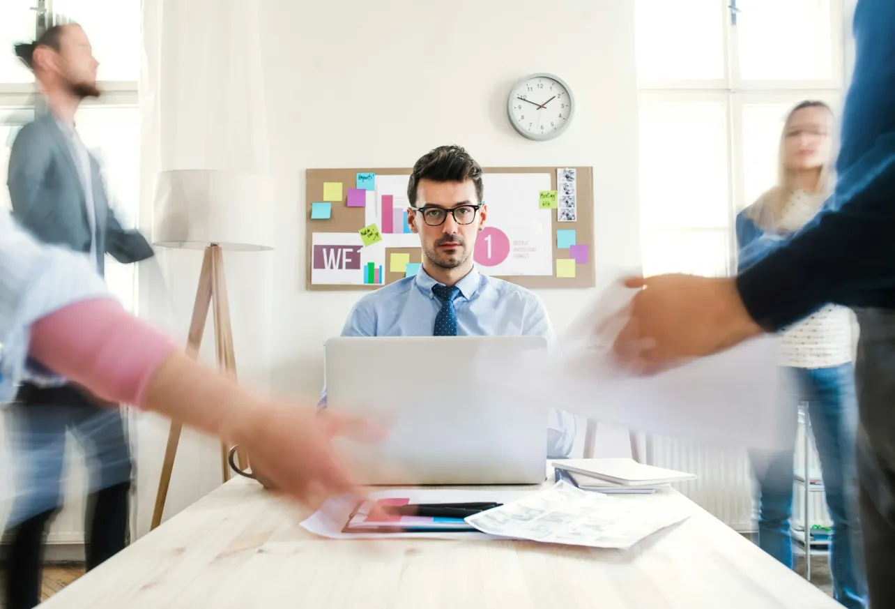 Office worker sitting at a laptop while blurred colleagues and papers move around him, suggesting workload pressure and work intensification.