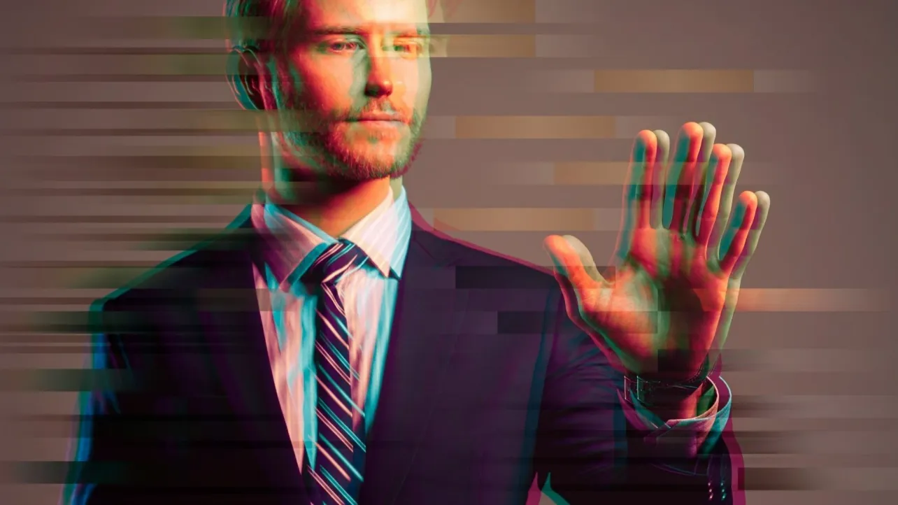 A suited professional holding up a hand in a stop gesture, with a glitch effect across the image, suggesting identity manipulation and digital deception in hiring.