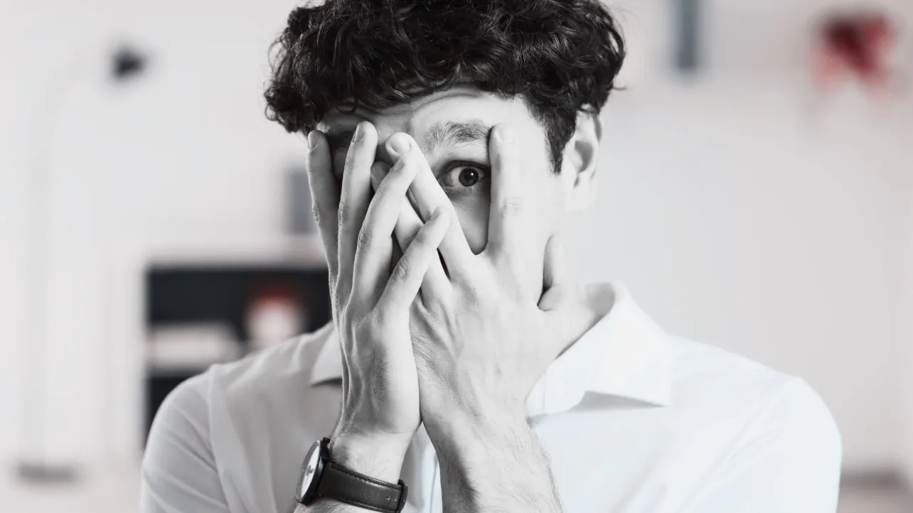 A young person covering most of their face with their hands, one eye visible, conveying anxiety, overwhelm, or apprehension in response to workplace change.