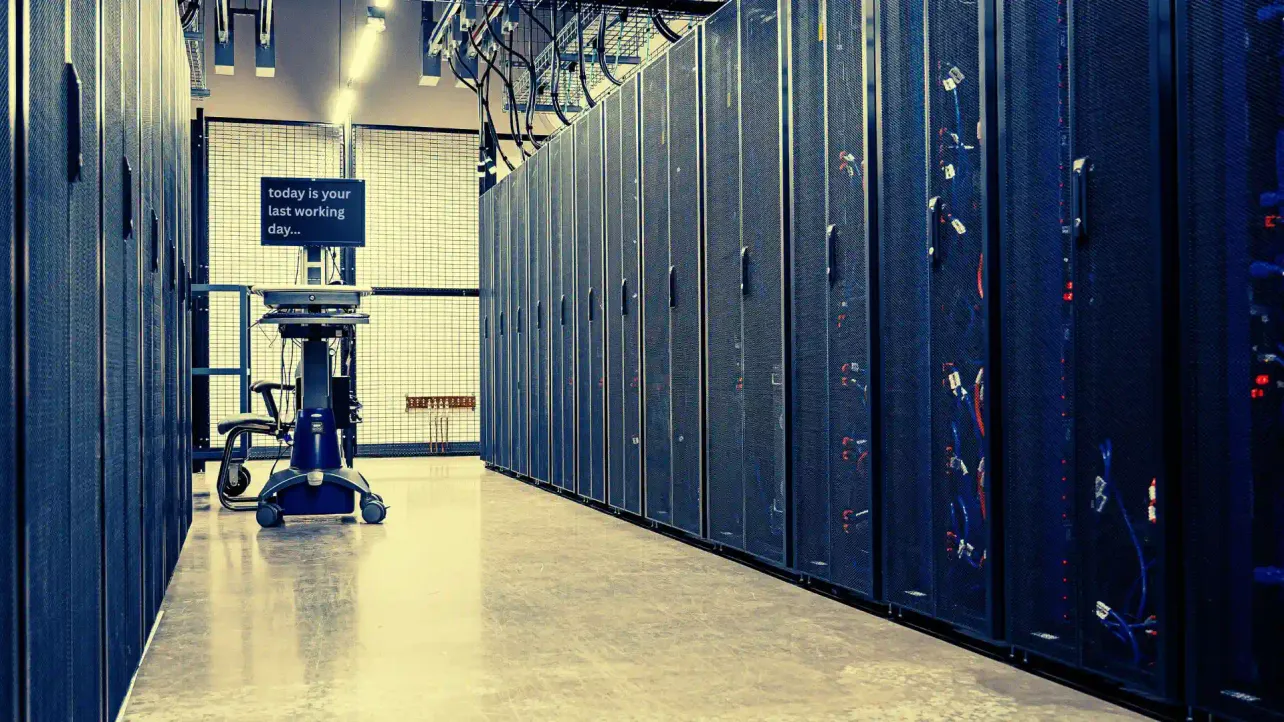 A data centre corridor with server racks on both sides and a workstation screen reading “today is your last working day…”, suggesting layoffs tied to infrastructure and AI spending.