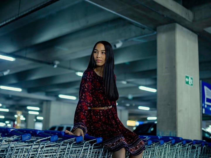 A young lady sits on a row of Ikea shopping carts in a concrete carpark, maybe contemplating her future career options